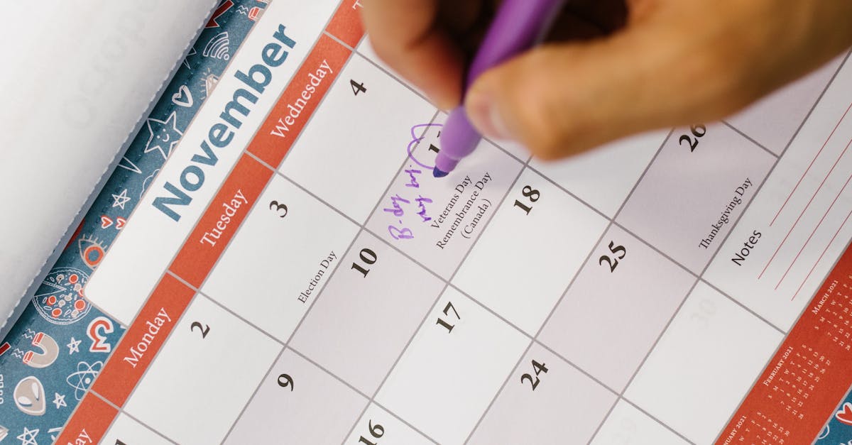 A person writes a reminder on a colorful November calendar using a purple pen.